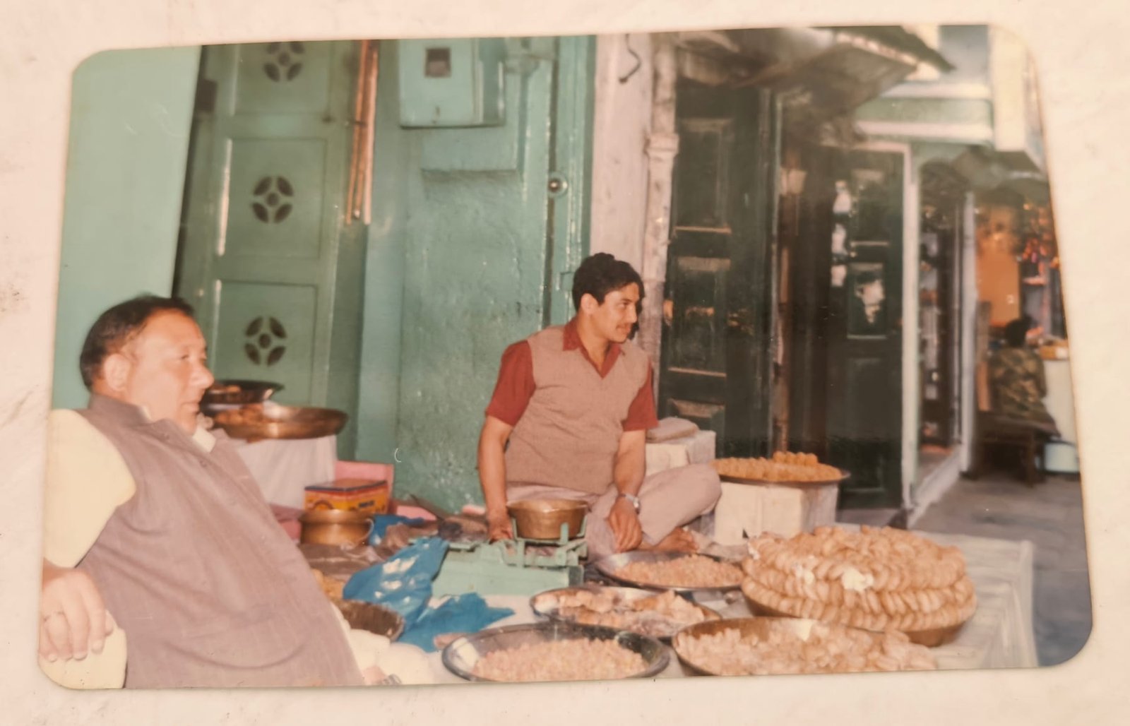 Archive photograph of a shopkeeper seated at the counter behind trays of freshly made sweets