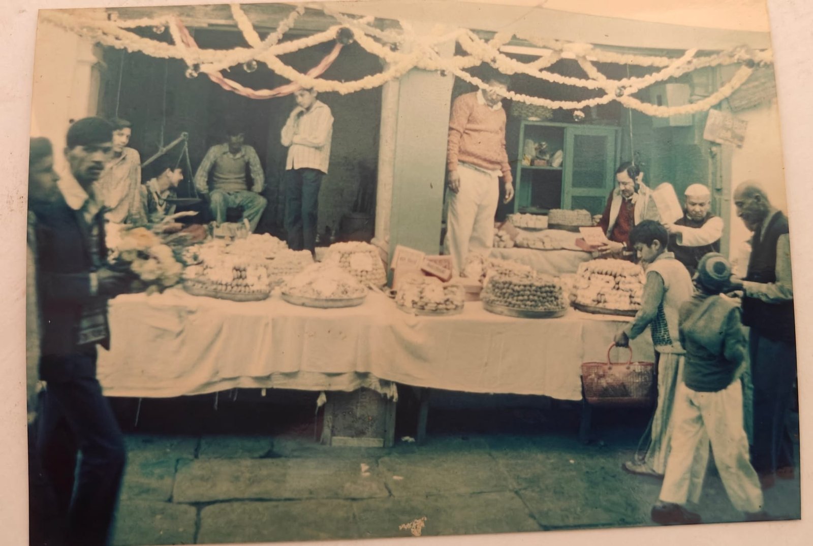 Faded archive photograph of the sweet stall dressed with garlands, trays of mithai laid out along a cloth-covered table